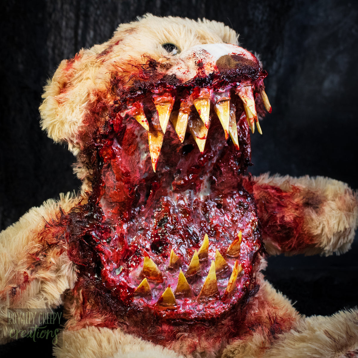 A horror prop of a bloody, screaming teddy bear with an open mouth showing sharp yellowed teeth and a gory red interior, photographed against a dark background.