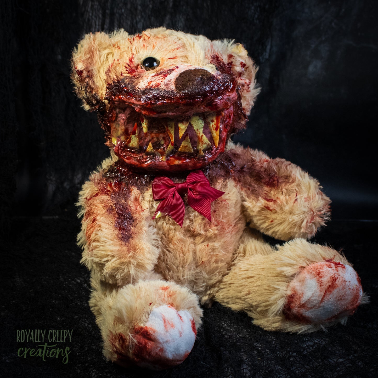 A horror-themed plush teddy bear with a beige fur body, bloody stitched details, a dark brown nose, a red bow, and a wide open mouth showing sharp teeth, posed against a dark textured background.