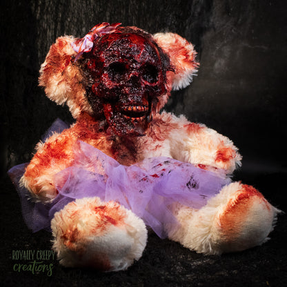 A horror-themed zombie teddy bear prop with a bloody, skull face, white fur stained with red blood, and a tattered lavender tutu, set against a dark textured background.