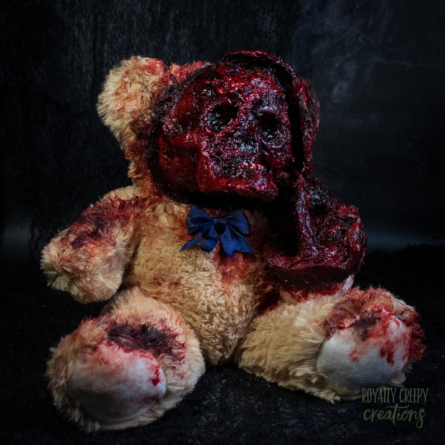 A horror-themed plush teddy bear with an exposed bloody skull for a face, and a dark blue bow tie, set against a dark textured background.