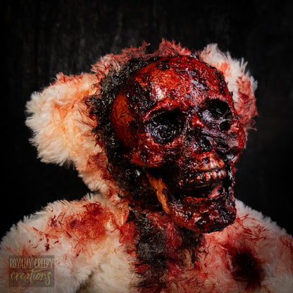 Close-up of a horror-themed plush teddy bear with an exposed bloody skull for a face, covered in fake blood splatters, set against a dark background.