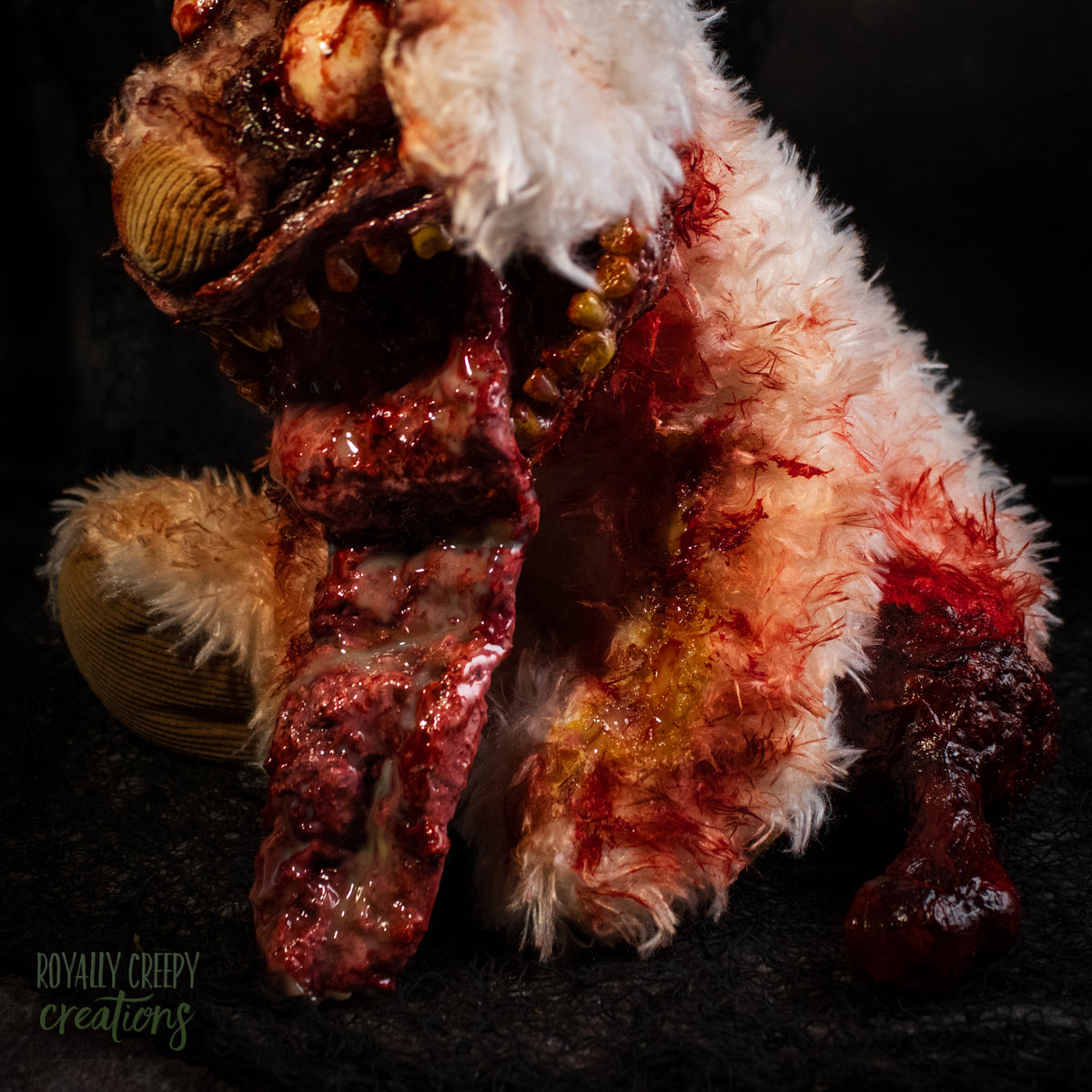 Side view of a horror-themed zombie dog plush toy with tattered fur, bloodshot red eyes, a bloody gaping mouth, and a protruding tongue, and a exposed bloody leg bone. Posed against a dark textured background.