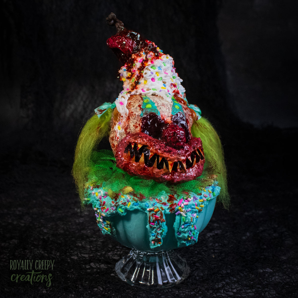 A highly detailed sculpture of a horror clown head staged like an ice cream sundae with a cherry on top it's head. The clown head is overflowing with whipped cream and ice cream and is sitting in a glass dessert bowl.