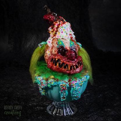A highly detailed sculpture of a horror clown head staged like an ice cream sundae with a cherry on top it's head. The clown head is overflowing with whipped cream and ice cream and is sitting in a glass dessert bowl.