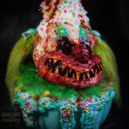 A close-up of a highly detailed sculpture of a horror clown head staged like an ice cream sundae. The clown head is overflowing with whipped cream and ice cream and is sitting in a glass dessert bowl.