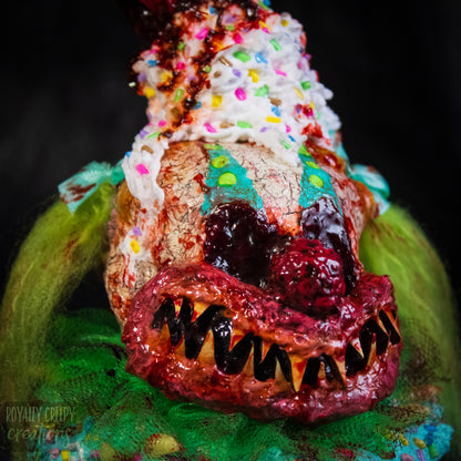 Close-up of a  highly detailed sculpture of a horror clown head staged like an ice cream sundae. The clown head is overflowing with whipped cream and ice cream and is sitting in a glass dessert bowl.