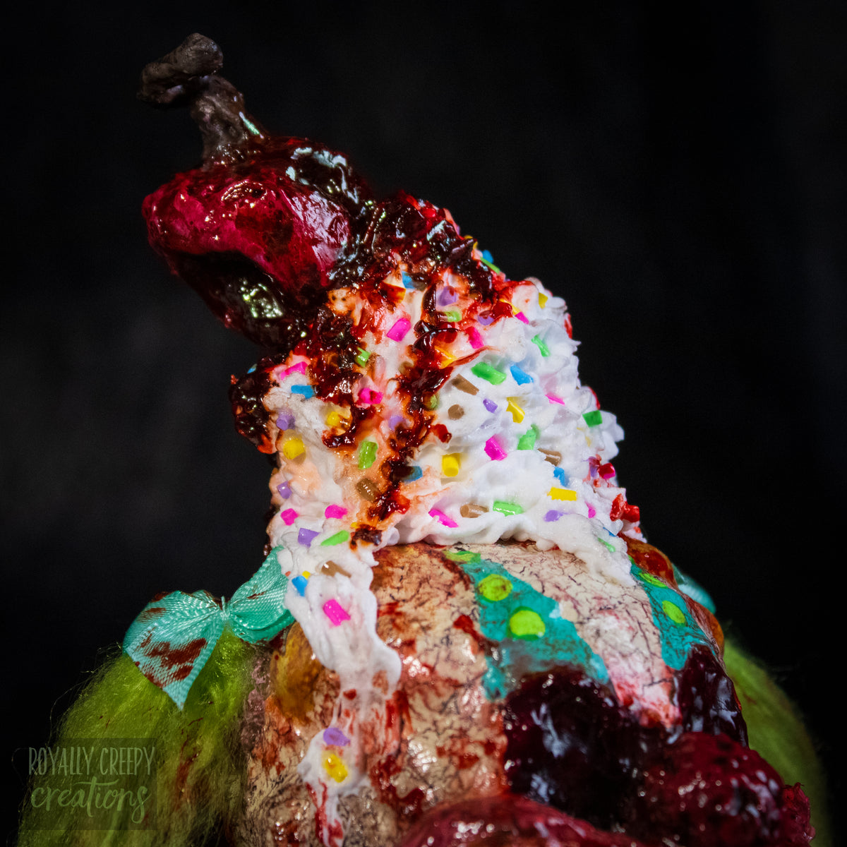 A Close-up of  highly detailed sculpture of a horror clown head staged like an ice cream sundae with a cherry and whipped cream with sprinkles on top it's head.