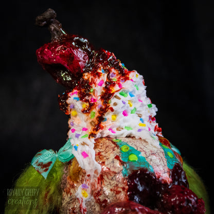 A Close-up of  highly detailed sculpture of a horror clown head staged like an ice cream sundae with a cherry and whipped cream with sprinkles on top it's head.