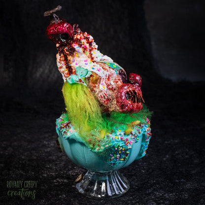 A side view of a highly detailed sculpture of a horror clown head staged like an ice cream sundae with a cherry on top it's head. The clown head is overflowing with whipped cream and ice cream and is sitting in a glass dessert bowl.