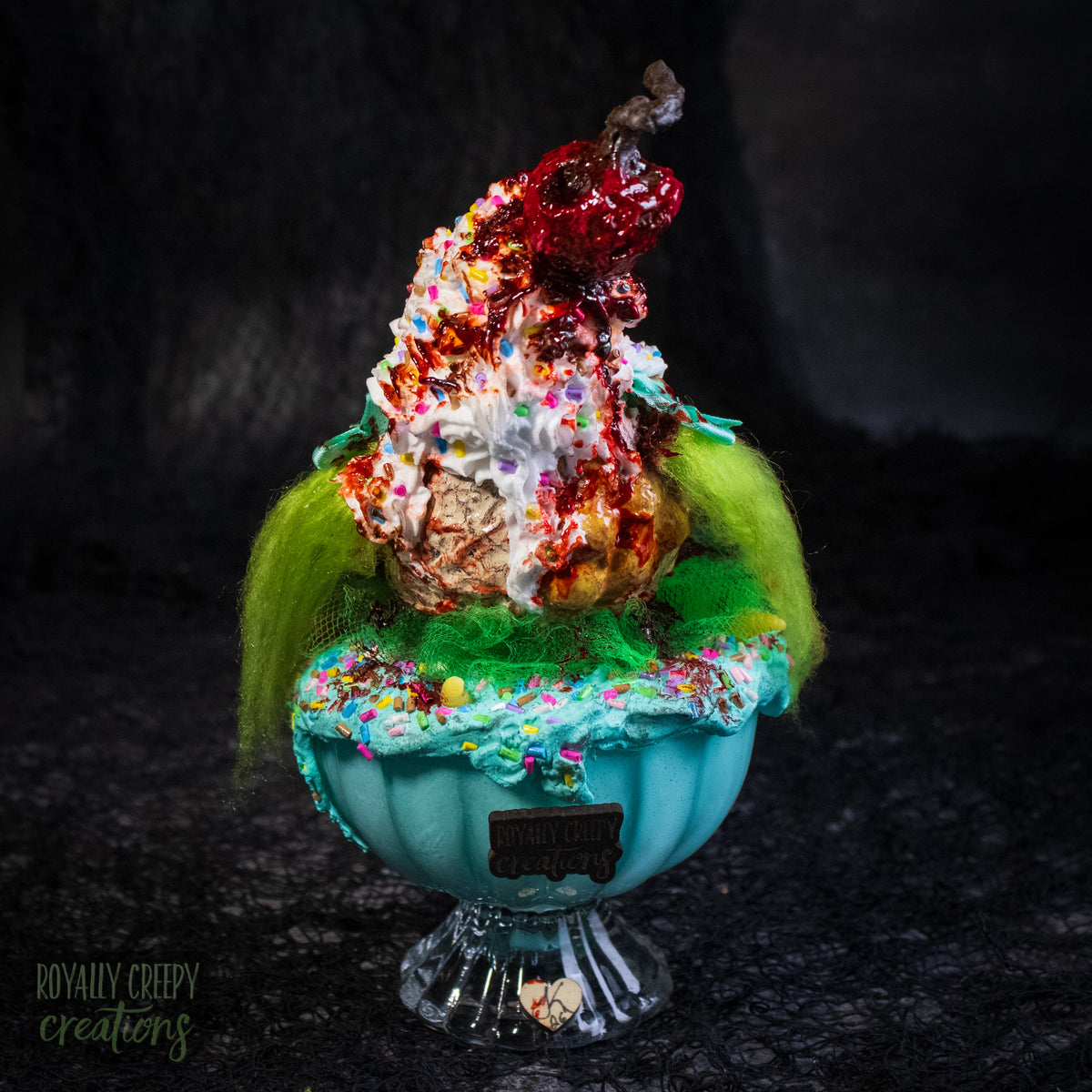 A back view of a highly detailed sculpture of a horror clown head staged like an ice cream sundae with a cherry on top it's head. The clown head is overflowing with whipped cream and ice cream and is sitting in a glass dessert bowl.
