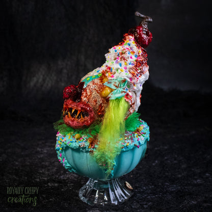 A side view of a highly detailed sculpture of a horror clown head staged like an ice cream sundae with a cherry on top it's head. The clown head is overflowing with whipped cream and ice cream and is sitting in a glass dessert bowl.