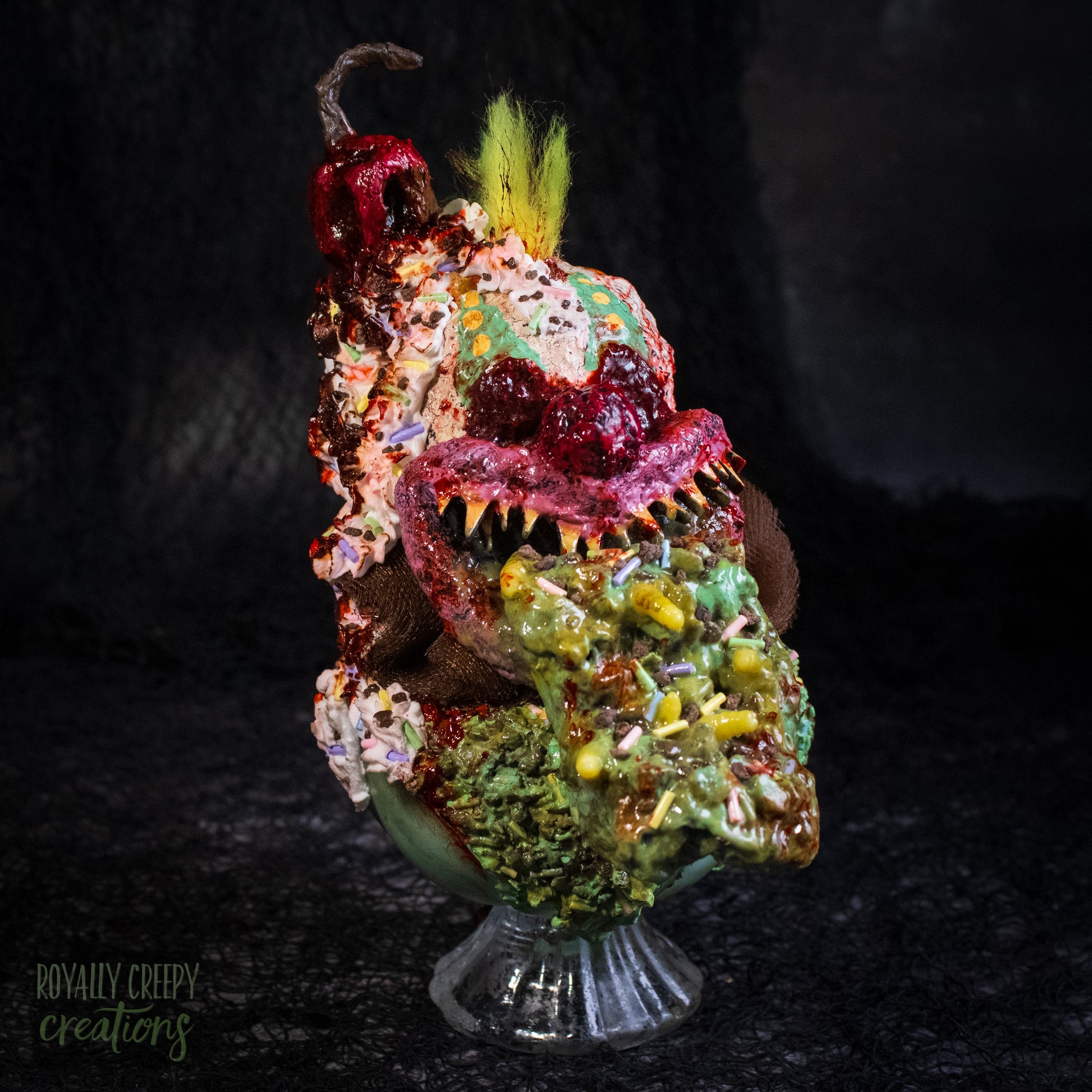 A highly detailed sculpture of a horror clown head staged like an ice cream sundae with a cherry on top it's head. The clown head is overflowing with whipped cream and ice cream and is sitting in a glass dessert bowl.