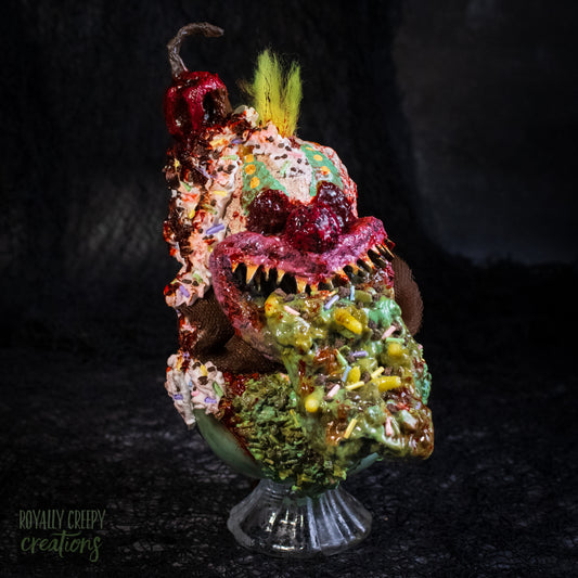 A highly detailed sculpture of a horror clown head staged like an ice cream sundae with a cherry on top it's head. The clown head is overflowing with whipped cream and ice cream and is sitting in a glass dessert bowl.