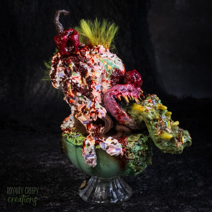 Side view of a highly detailed sculpture of a horror clown head staged like an ice cream sundae with a cherry on top it's head. The clown head is overflowing with whipped cream and ice cream and is sitting in a glass dessert bowl.