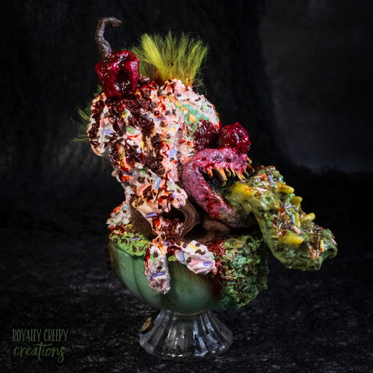 Side view of a highly detailed sculpture of a horror clown head staged like an ice cream sundae with a cherry on top it's head. The clown head is overflowing with whipped cream and ice cream and is sitting in a glass dessert bowl.