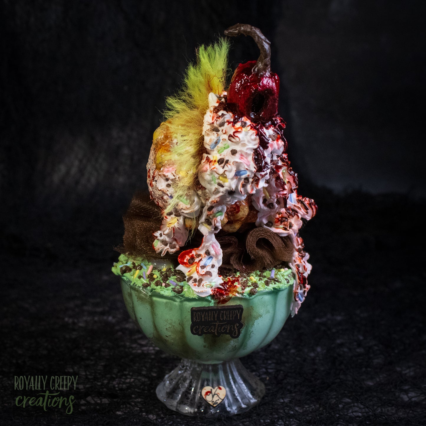 Back view of a highly detailed sculpture of a horror clown head staged like an ice cream sundae with a cherry on top it's head. The clown head is overflowing with whipped cream and ice cream and is sitting in a glass dessert bowl.