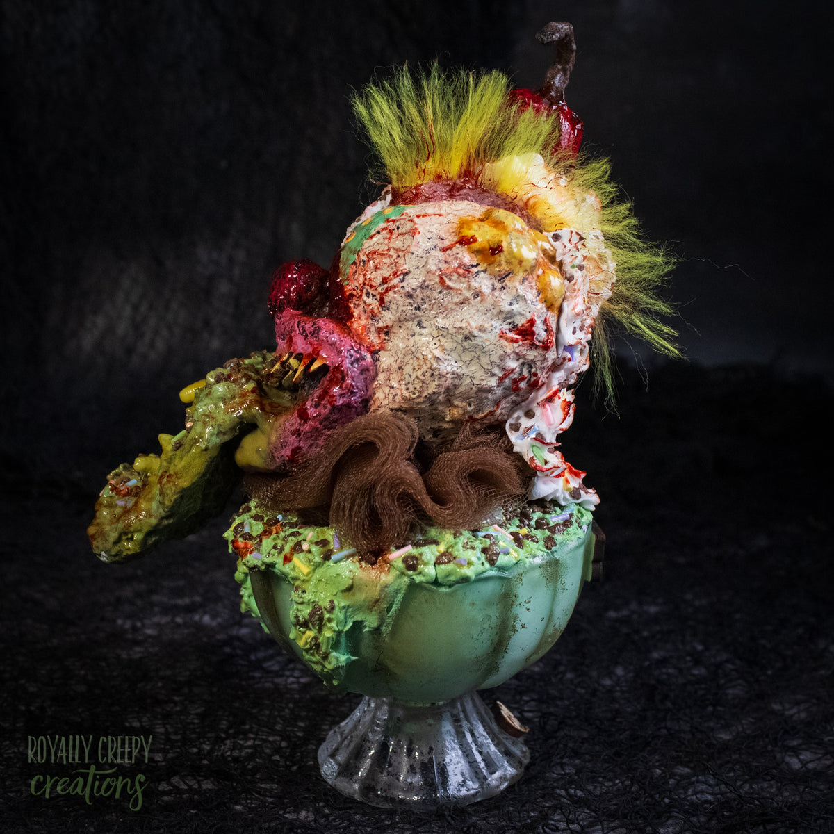 Side view of a highly detailed sculpture of a horror clown head staged like an ice cream sundae with a cherry on top it's head. The clown head is overflowing with whipped cream and ice cream and is sitting in a glass dessert bowl.