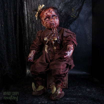 A creepy, horror-themed doll seated in a tattered brown outfit, covered in fake blood, with a bloody stitched mouth with drool and a bow in its hair, set against a dark textured background.