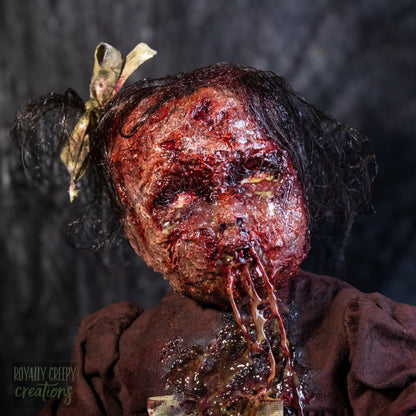 A close-up of a creepy, horror-themed doll seated covered in fake blood, with a bloody stitched mouth with drool and a bow in its hair, set against a dark textured background.