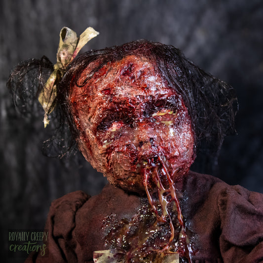 A close-up of a creepy, horror-themed doll seated covered in fake blood, with a bloody stitched mouth with drool and a bow in its hair, set against a dark textured background.