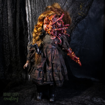 A creepy horror doll, wearing a dark distressed dress and boots, with long golden-brown hair and a grotesque red-orange fleshy appendage replacing half the face, against a dark textured backdrop. 
