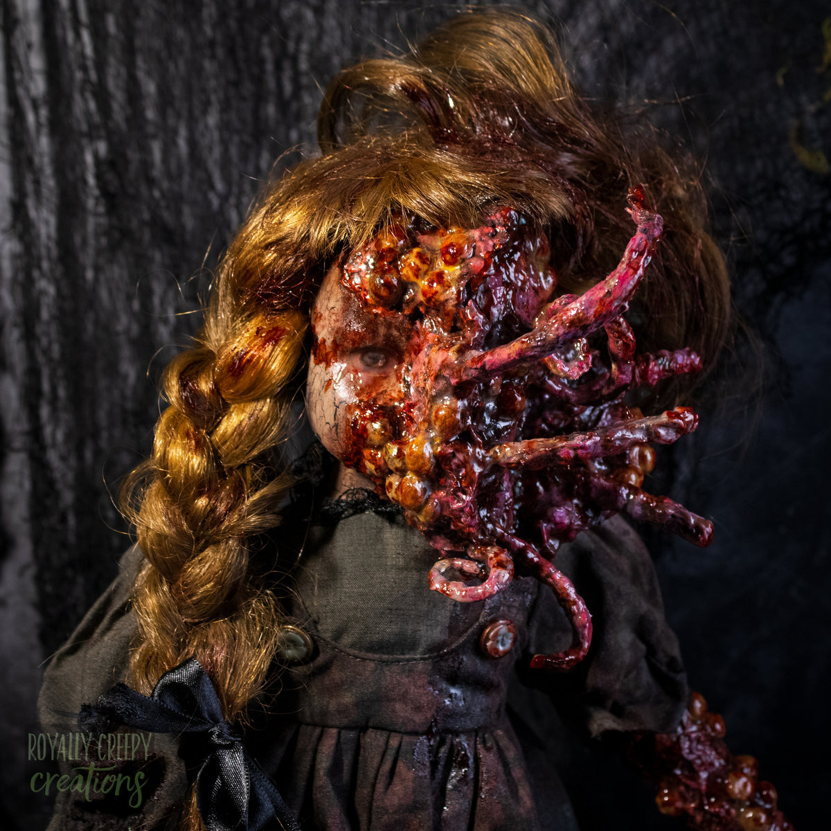 A close-up of a creepy horror doll, with long golden-brown hair and a grotesque red-orange fleshy appendage replacing half the face, against a dark textured backdrop. 