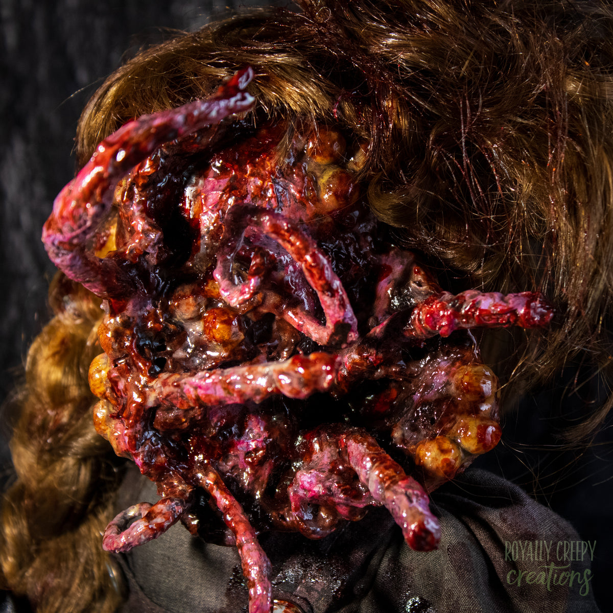 A close-up of a creepy horror doll, with long golden-brown hair and a grotesque red-orange fleshy appendage replacing half the face, against a dark textured backdrop. 