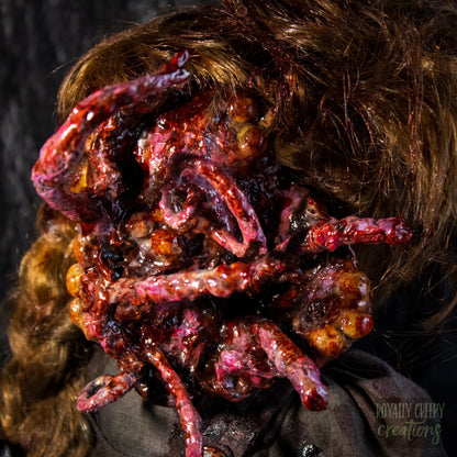 A close-up of a creepy horror doll, with long golden-brown hair and a grotesque red-orange fleshy appendage replacing half the face, against a dark textured backdrop. 