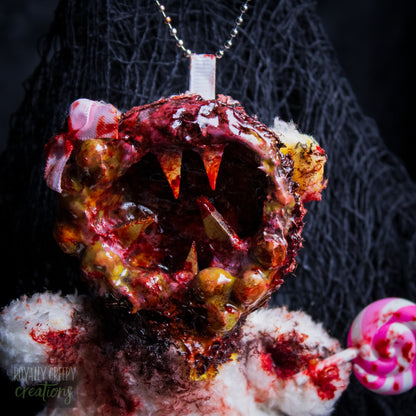 A close-up of a horror-themed plush bear keychain with a bloody, gory mouth design, fuzzy body, and lollipop accessory, shown against a black netted backdrop.