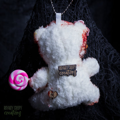 A white fluffy teddy bear plush pendant or keychain with a silver chain, featuring a 'Royally Creepy Creations' metal pin on the chest, a small heart charm, and a pink and white lollipop accessory, set against a dark textured background.