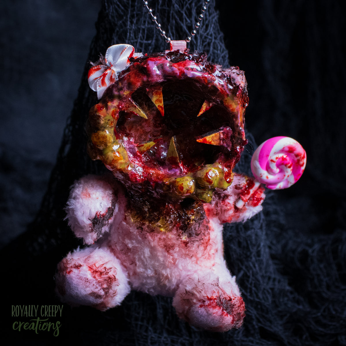 A horror-themed plush bear keychain with a bloody, gory mouth design, fuzzy body, and lollipop accessory, shown against a black netted backdrop.