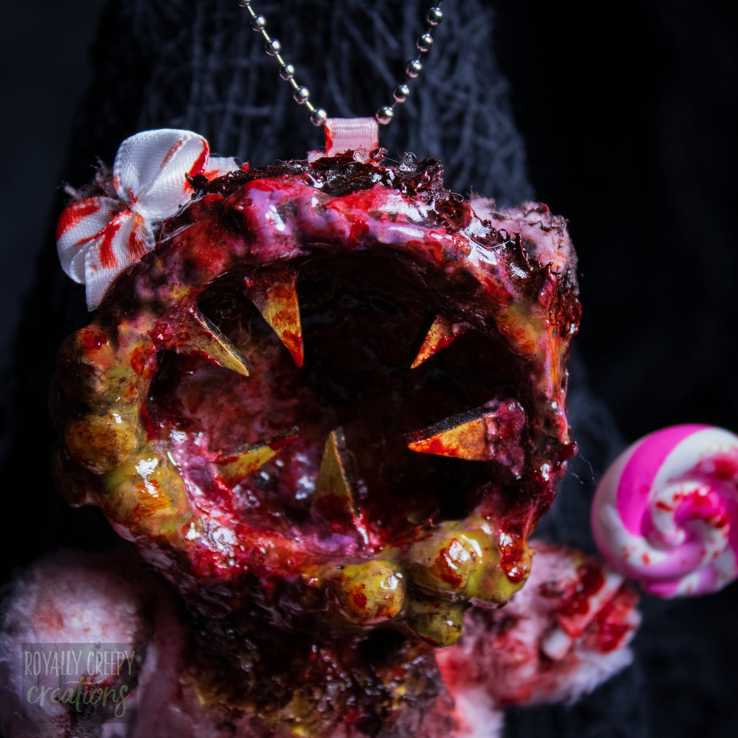 A close-up of a horror-themed plush bear keychain with a bloody, gory mouth design, fuzzy body, and lollipop accessory, shown against a black netted backdrop.