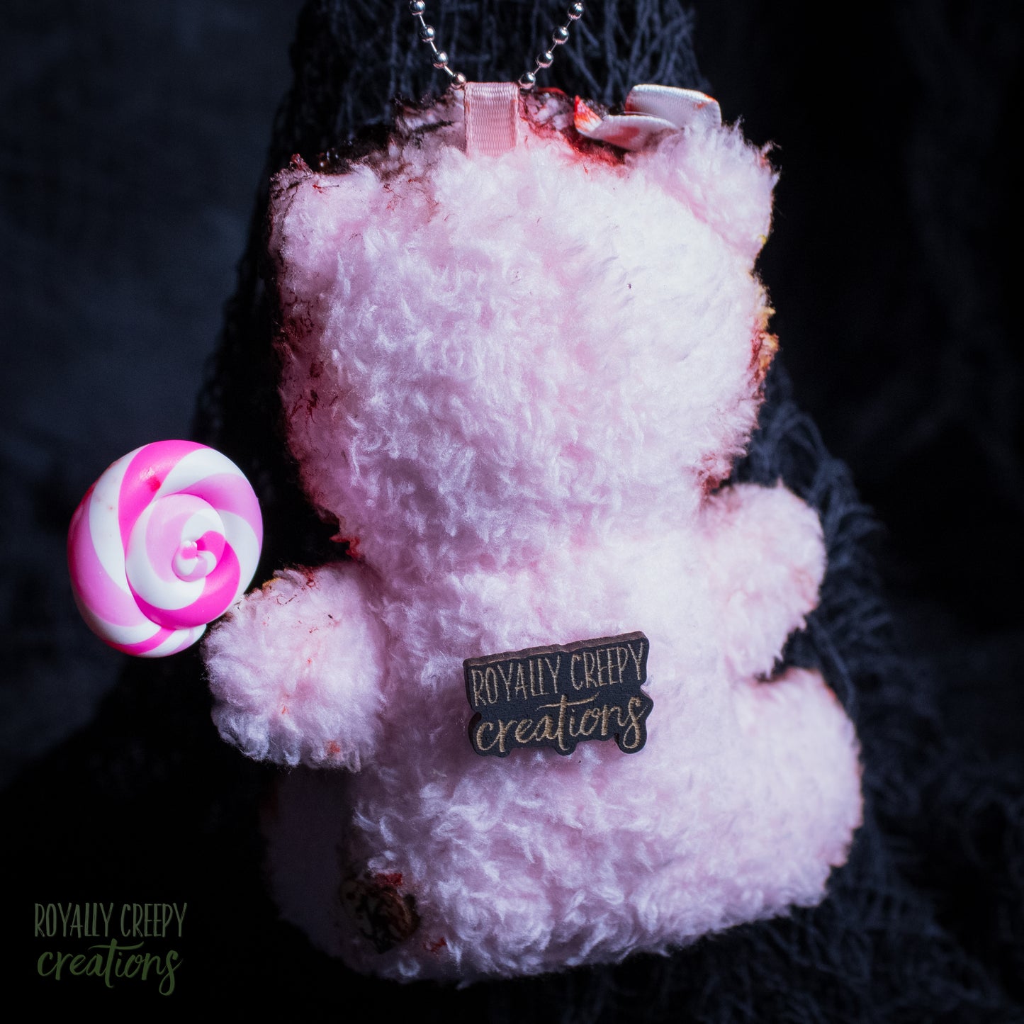 A fluffy light pink teddy bear keychain or charm with a metal chain, featuring a small black pin that reads 'Royally Creepy Creations' and holding a pink and white swirl lollipop, set against a dark textured background.