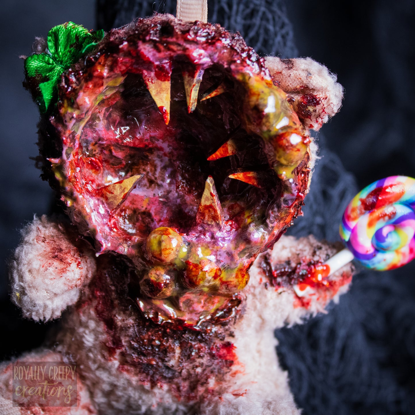 A close-up of a horror-themed plush bear keychain with a bloody, gory mouth design, fuzzy body, and lollipop accessory, shown against a black netted backdrop.