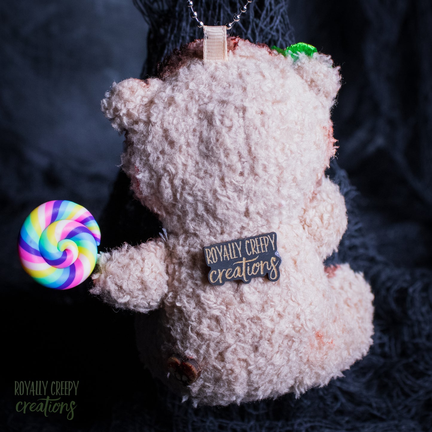 A plush teddy bear ornament or keychain in a shaggy texture, shown from the back with a colorful rainbow lollipop accessory attached on the left. The bear has a tag reading 'ROYALLY CREEPY creations' on its back, photographed against a dark textured background.