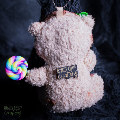 A plush teddy bear ornament or keychain in a shaggy texture, shown from the back with a colorful rainbow lollipop accessory attached on the left. The bear has a tag reading 'ROYALLY CREEPY creations' on its back, photographed against a dark textured background.