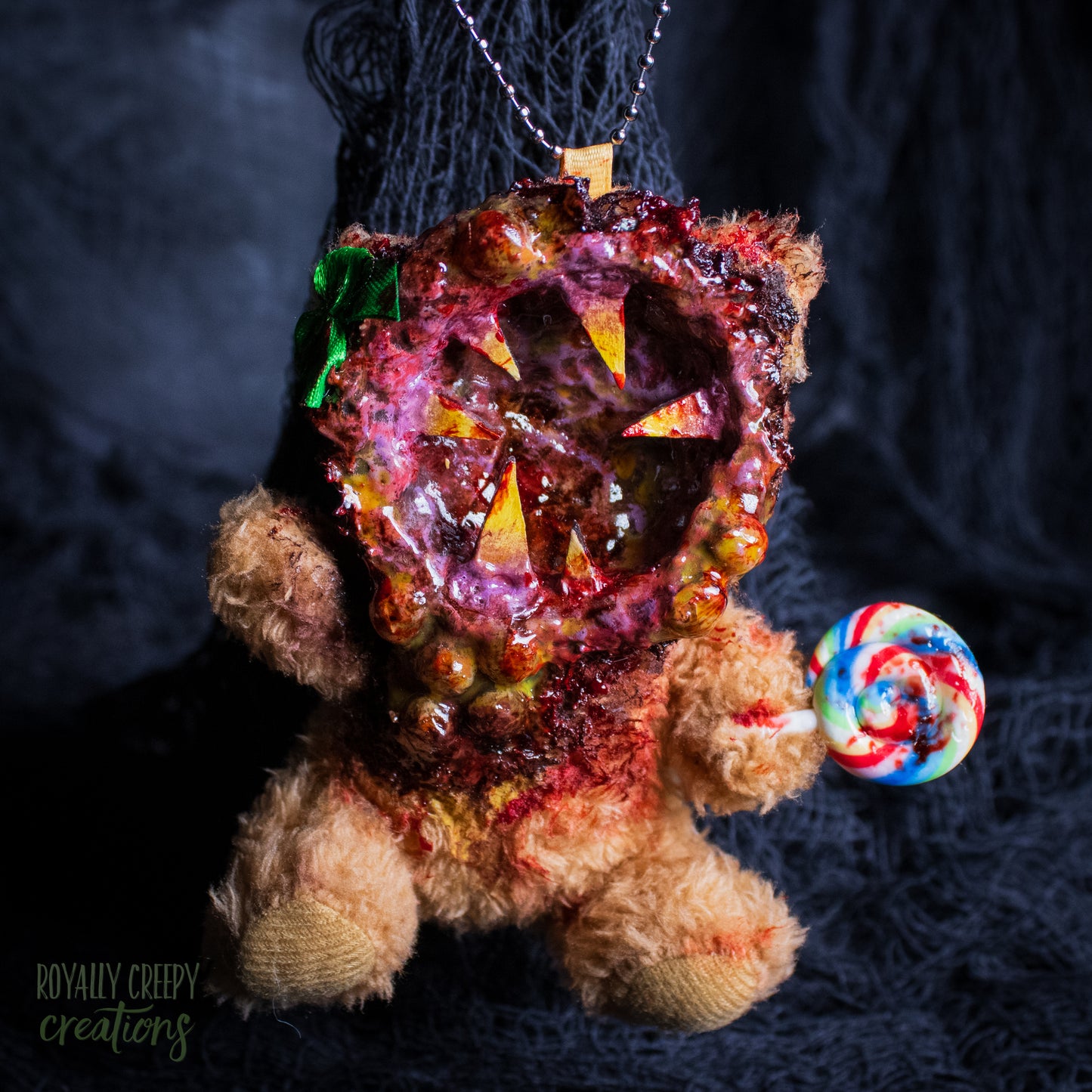 A horror-themed plush bear keychain with a bloody, gory mouth design, fuzzy body, and lollipop accessory, shown against a black netted backdrop.