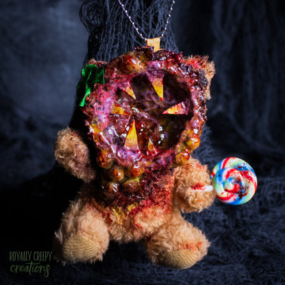 A horror-themed plush bear keychain with a bloody, gory mouth design, fuzzy body, and lollipop accessory, shown against a black netted backdrop.