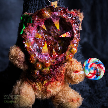 A close-up of a horror-themed plush bear keychain with a bloody, gory mouth design, fuzzy body, and lollipop accessory, shown against a black netted backdrop.