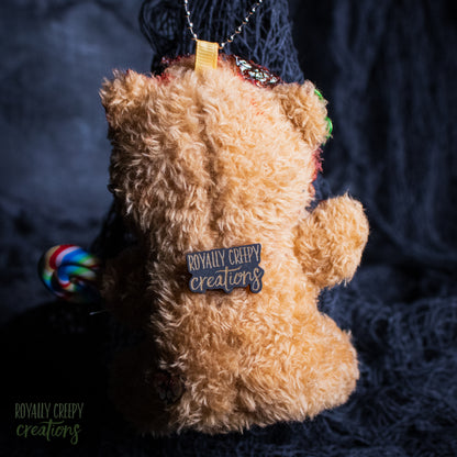 A plush teddy bear ornament or keychain shown from the back, featuring a tan fuzzy body, a pin reading 'ROYALLY CREEPY creations' on the back, a colorful lollipop decoration on the left, and a hanging loop at the top, set against a dark textured fabric background.