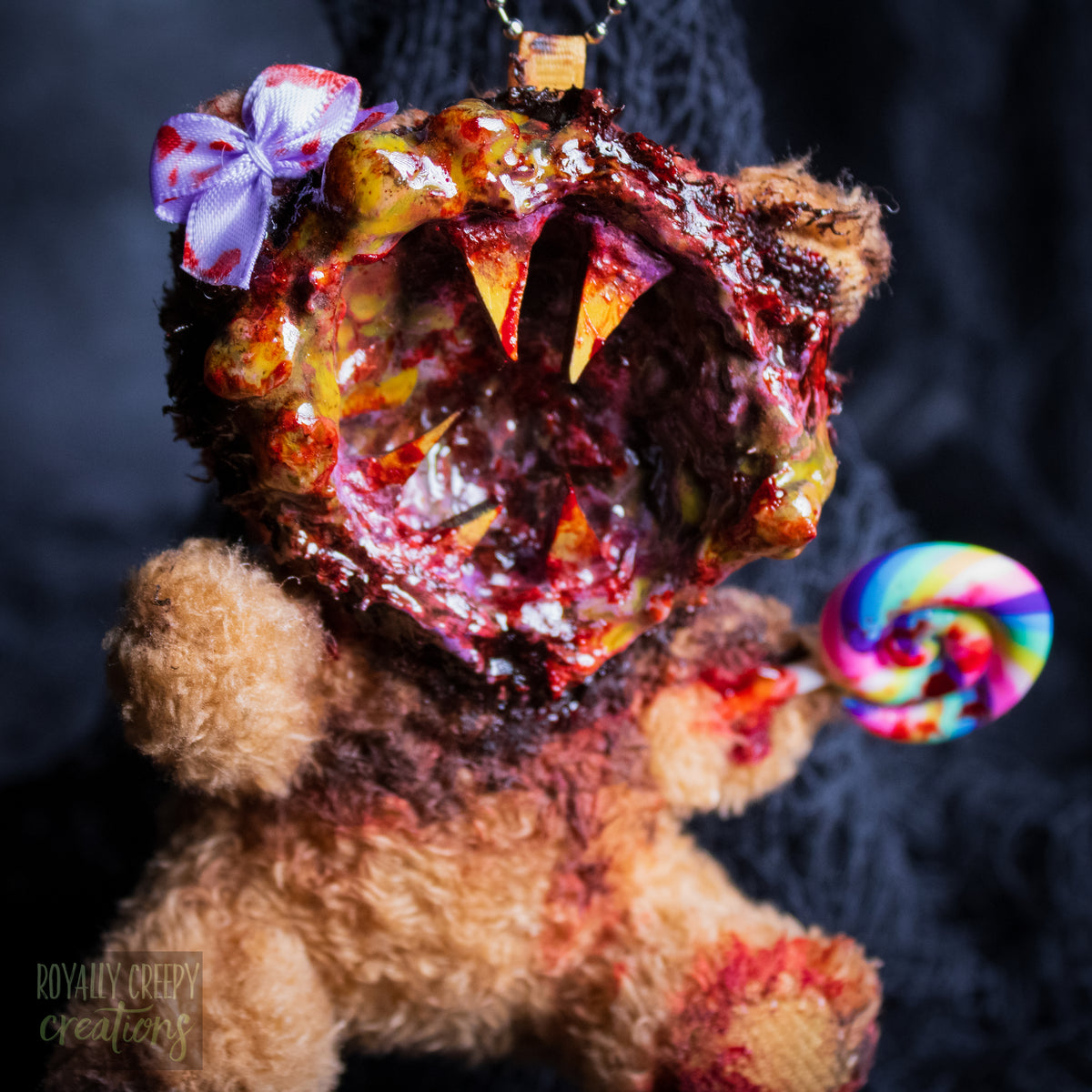 A close-up of a horror-themed plush bear keychain with a bloody, gory mouth design, fuzzy body, and lollipop accessory, shown against a black netted backdrop.