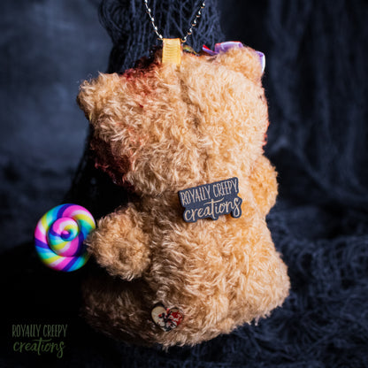 A plush teddy bear ornament in light brown fur, holding a rainbow spiral lollipop and wearing a black patch that reads 'ROYALLY CREEPY creations'. The ornament hangs from a silver chain against a dark textured background.