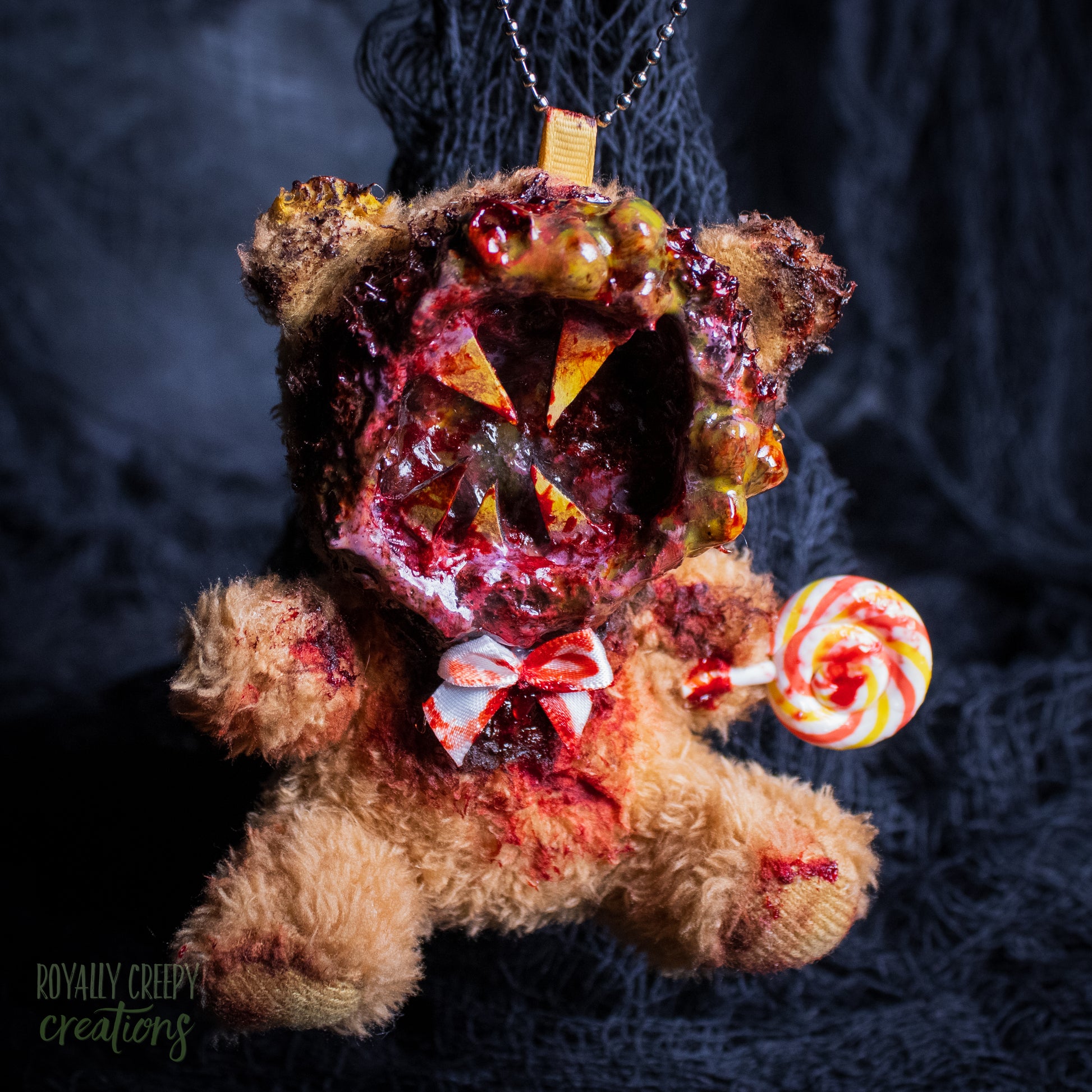 A horror-themed plush bear keychain with a bloody, gory mouth design, fuzzy body, and lollipop accessory, shown against a black netted backdrop.