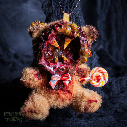 A horror-themed plush bear keychain with a bloody, gory mouth design, fuzzy body, and lollipop accessory, shown against a black netted backdrop.