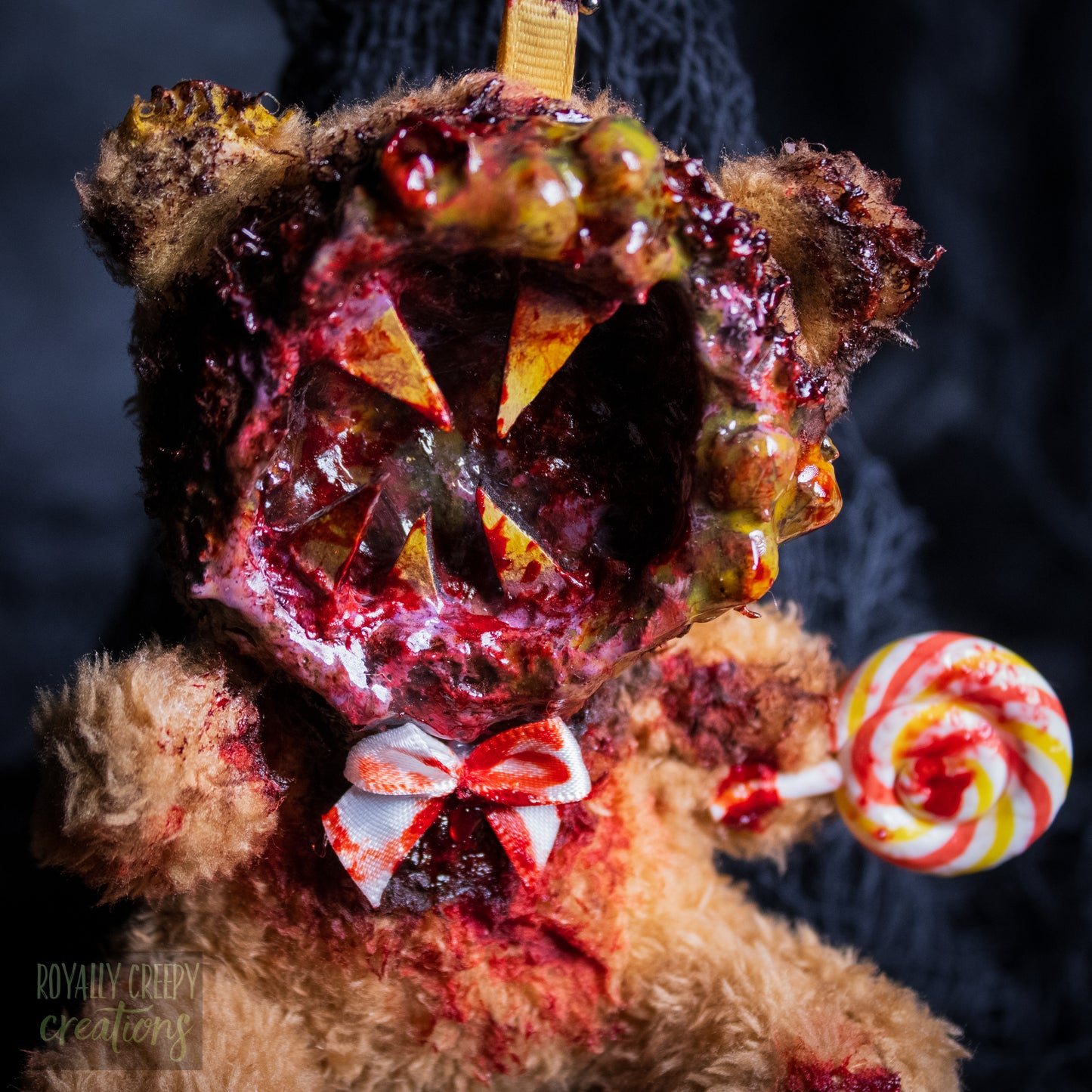 A close-up of a horror-themed plush bear keychain with a bloody, gory mouth design, fuzzy body, and lollipop accessory, shown against a black netted backdrop.