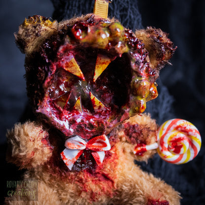 A close-up of a horror-themed plush bear keychain with a bloody, gory mouth design, fuzzy body, and lollipop accessory, shown against a black netted backdrop.