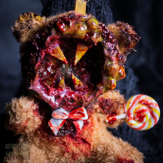 A close-up of a horror-themed plush bear keychain with a bloody, gory mouth design, fuzzy body, and lollipop accessory, shown against a black netted backdrop.