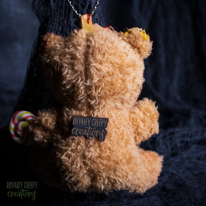 A plush teddy bear ornament shown from the back, made of curly tan faux fur with a small black tag reading 'ROYALLY CREEPY creations' and a silver hanging loop. A small lollipop decoration is attached on the left side, set against a dark textured fabric background.