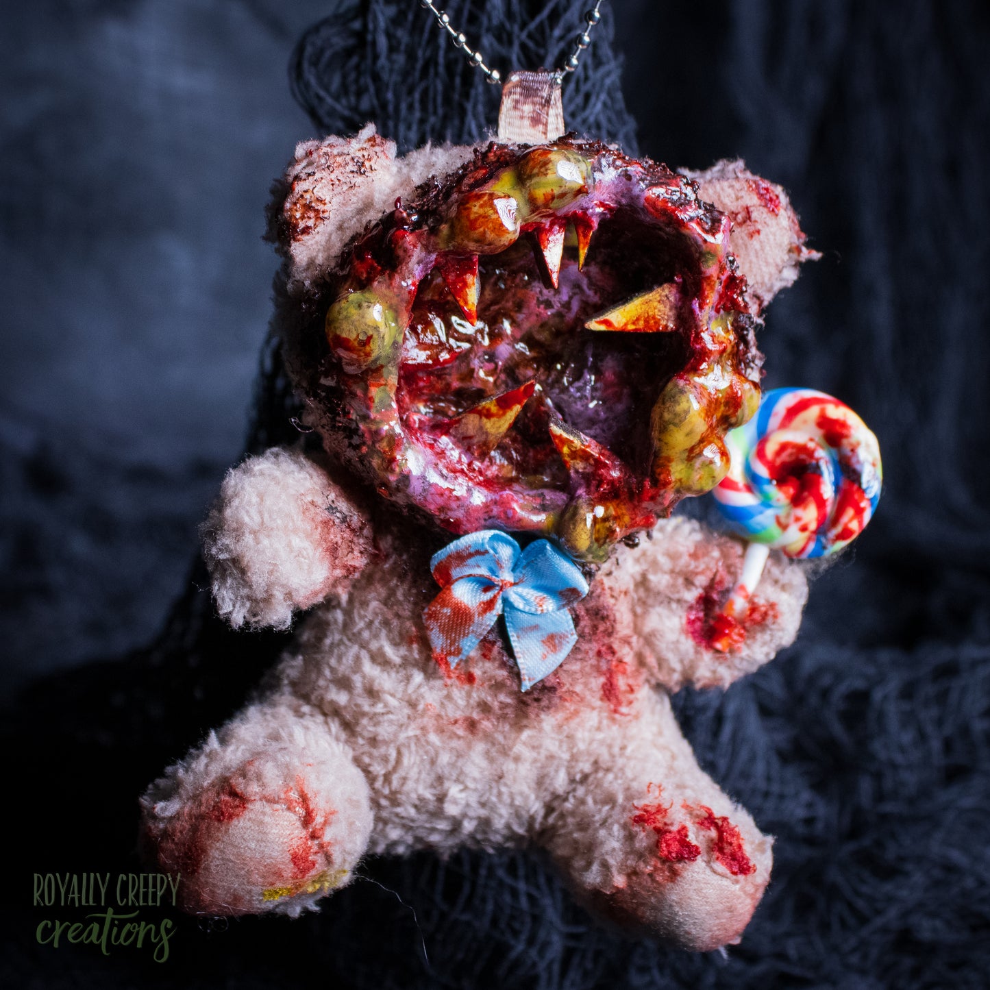 A horror-themed plush bear keychain with a bloody, gory mouth design, fuzzy body, and lollipop accessory, shown against a black netted backdrop.