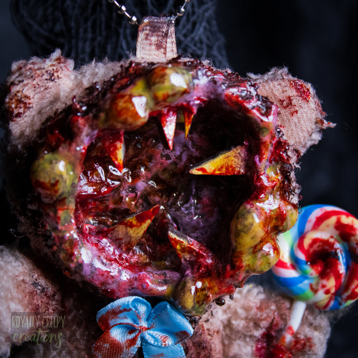 A close-up of a horror-themed plush bear keychain with a bloody, gory mouth design, fuzzy body, and lollipop accessory, shown against a black netted backdrop.